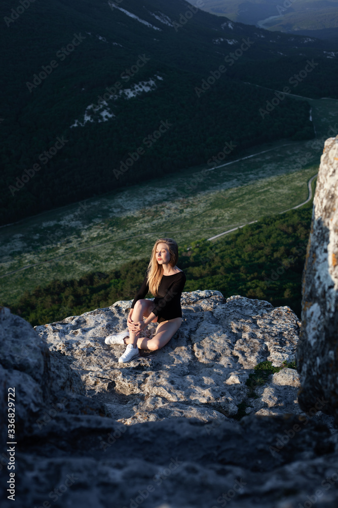 Beautiful young girl sitting on a rock. Antistress and mindfullness in a moment. Incredible view of mountains with green trees. Sunset time. Place for your text.