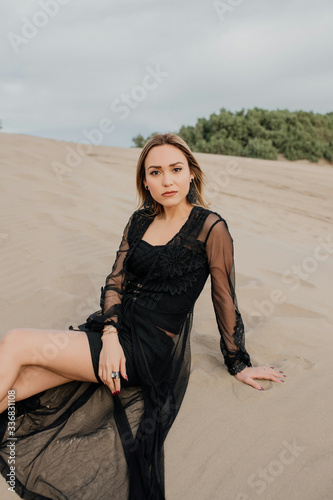 Mujer elegante con vestido negro sentada sobre la arena en la playa mirando directamente a la cámara photo