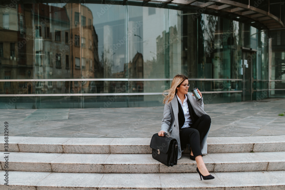 Fototapeta premium A young attractive businesswoman with a bag is sitting on the stairs in front of the company