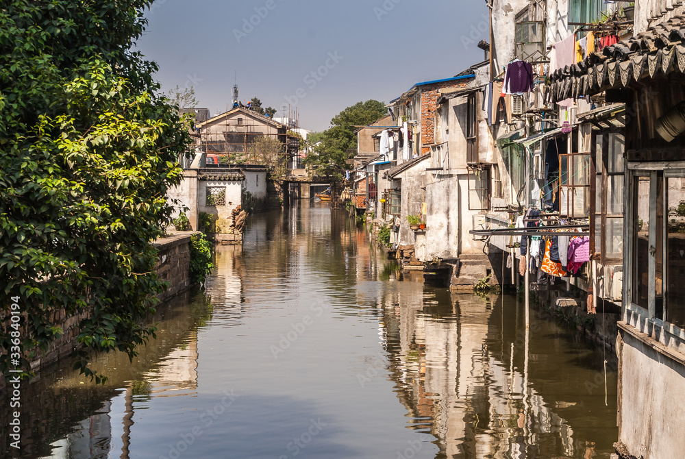 Suzhou China - May 3, 2010: City canal with brown water reflects the backside of houses along the water, and green foliage on other side under blue sky. Laundry add colors.
