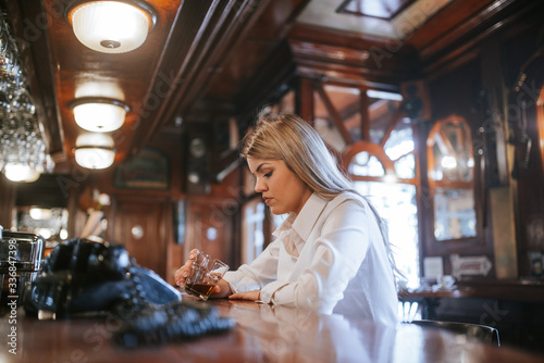 A young attractive caucasian blonde girl with a drink in her hand sits at a bar