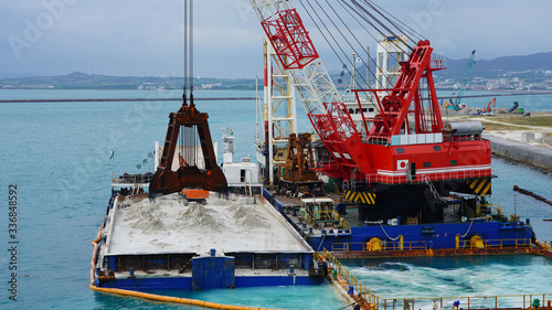Huge excavator on floating platform in the blue water of the Pacific Ocean extract white sand from the bottom and load onto a barge amid clouds and tropical islands. close up crane basket plunge