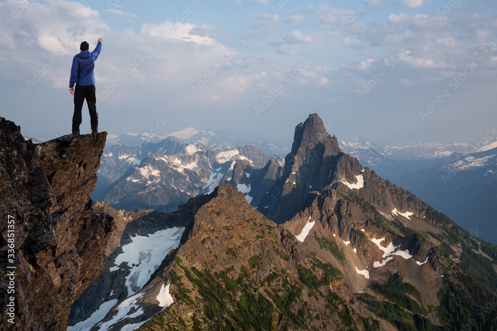 Adventurous Man Hiker With Hands Up on top of a Steep Rocky Cliff. Sunset or Sunrise. Composite. Landscape Taken from British Columbia, Canada. Concept: Adventure, Explore, Hike, Lifestyle