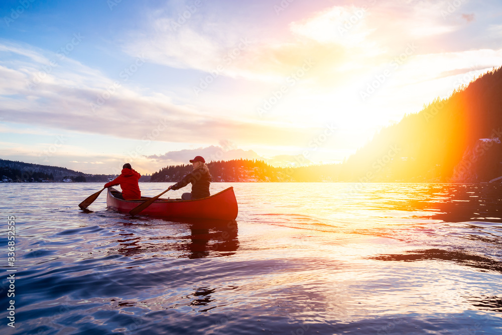 Couple friends on a wooden canoe are paddling in water during a vibrant sunny sunset. Taken in Indian Arm, near Deep Cove, North Vancouver, British Columbia, Canada. Concept: Adventure, Sport, Explore