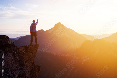 Adventurous Man Hiker With Hands Up on top of a Steep Rocky Cliff. Sunset or Sunrise. Composite. Landscape Taken from British Columbia, Canada. Concept: Adventure, Explore, Hike, Lifestyle