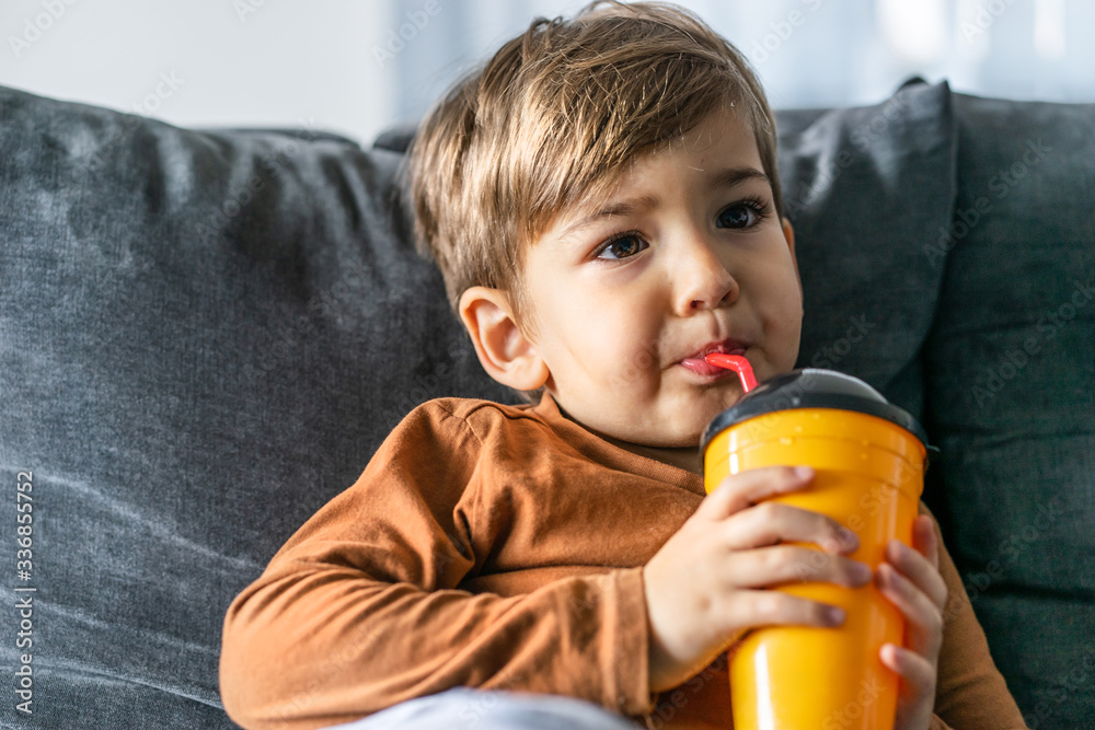 Portrait of small cute caucasian boy three or four years old sitting on the bed or sofa at home holding a plastic cup with straw drinking juice or soda at home alone in day looking to side half length Portrait of small cute caucasian boy three or four years old sitting on the bed or sofa at home holding a plastic cup with straw drinking juice or soda at home alone in day looking to side half length