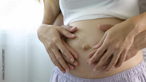 Wallpaper Mural Young couple expecting baby standing together indoors. Pregnant woman hand of husband hugging the tummy in white room near window light. Torontodigital.ca