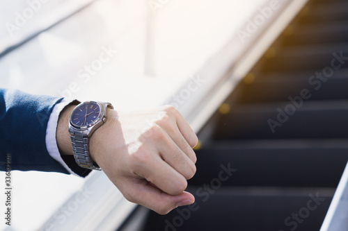 The photo close up of businessman watching his watch in his hand while standing on the escalator.  Business and finance concept.