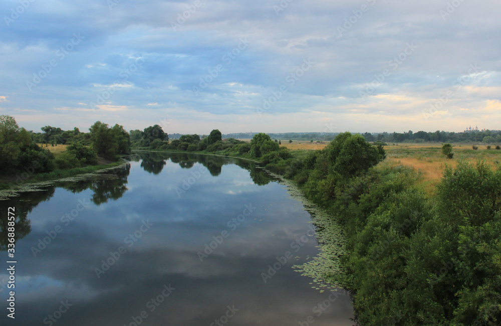 A wide river at a bend surrounded by trees and bushes. Blue clouds are reflected in the surface of the river.