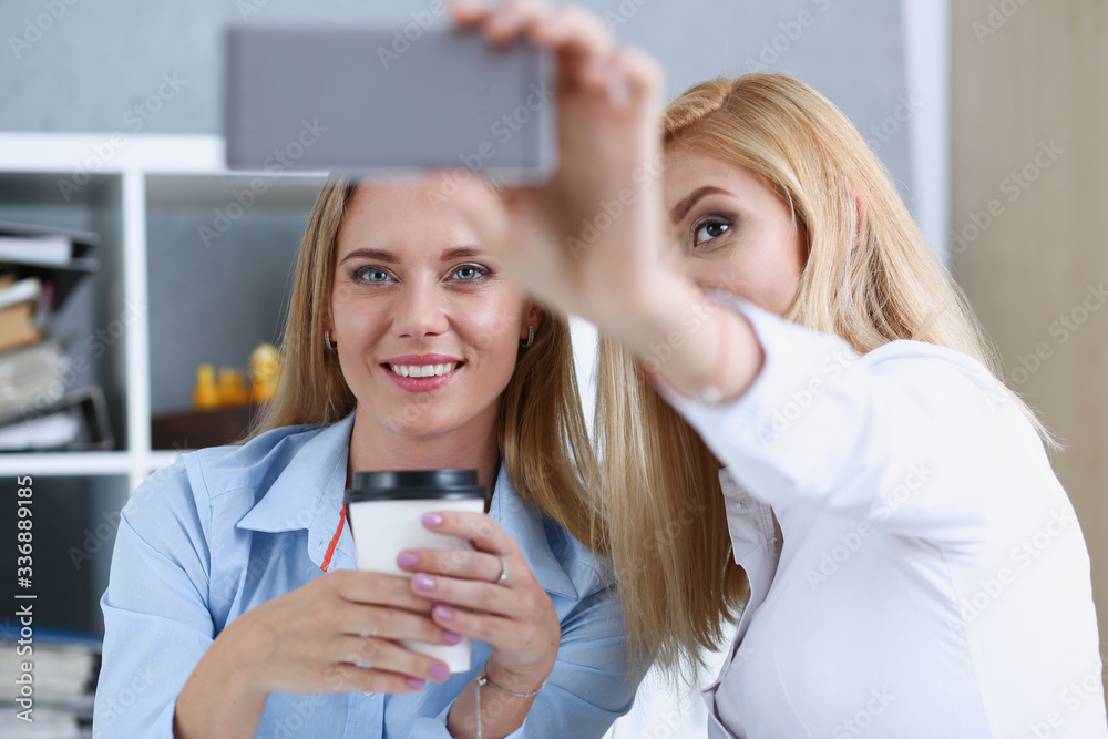 Two businesswomen in the office smile and do selfie on a smartphone during a break to have coffee Two businesswomen in the office smile and do selfie on a smartphone during a break to have coffee