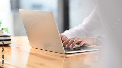 Cropped image of creative woman's hands typing on computer laptop that putting on wooden working desk and surrounded by stack of books, pencil holder, coffee cup and potted plant. Orderly workspace.