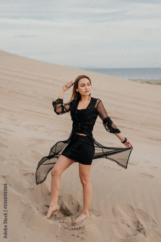 Foto de Mujer rubia y elegante mirando hacia la playa , parada sobre la  arena, con vestido negro y descalza. do Stock