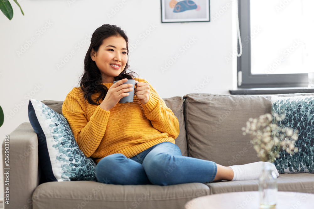 people and leisure concept - happy smiling asian young woman in yellow sweater sitting on sofa and drinking coffee at home