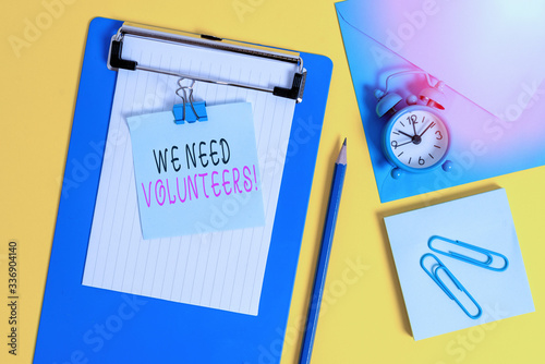 Word writing text We Need Volunteers. Business photo showcasing someone who does work without being paid for it Clipboard sheet note binder pencil clips clock envelop colored background photo