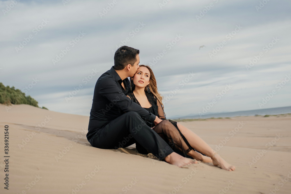 Pareja enamorados hombre besando a mujer en la cabeza , sentados en la playa con el mar y el cielo de fondo, vestidos de negro. 