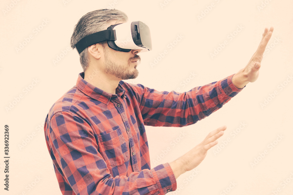 Focused man in VR headset touching air. Serious bearded man in checkered shirt moving with hands and using VR headset on grey background. Technology concept