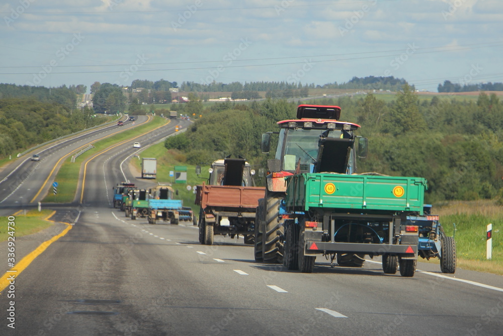 Tractors with flatbed trailers rides on asphalt suburban highway road on a Sunny summer day, agricultural mechanization, rear side view in perspective