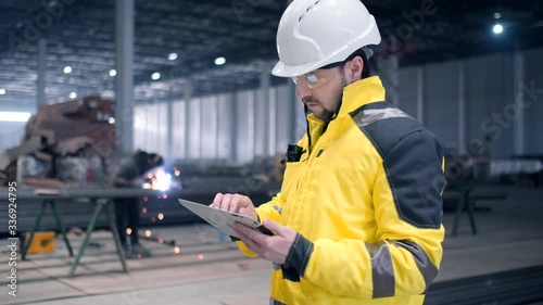 Portrait of Professional Heavy Industry Engineer / Worker Wearing Safety Uniform, Goggles and Hard Hat In the Background Unfocused Large Industrial Factory where Welding Sparks Flying. Safety glasses
