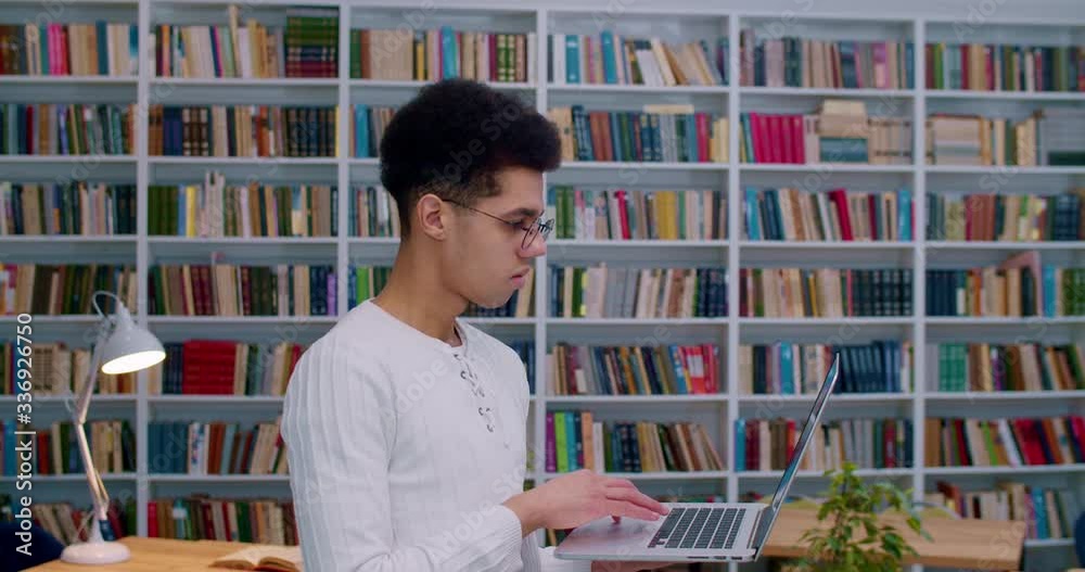 Male student in glasses holding laptop computer and typing on keyboard in library room. Latino guy turning face to camera and smiling cheerfully in bibliotheca while studying. Male student in glasses holding laptop computer and typing on keyboard in library room. Latino guy turning face to camera and smiling cheerfully in bibliotheca while studying.