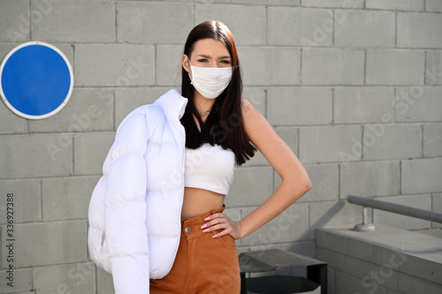 Outdoor photo of a young brunette girl in a medical mask in beige trousers and a white jacket on the background of the railing of a building s staircase in the city. Made on a sunny spring day. photo