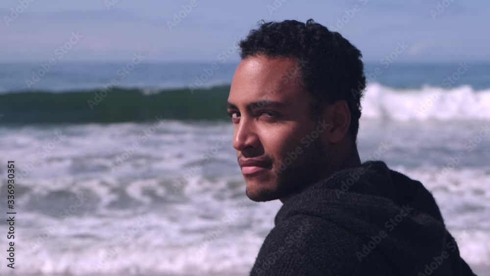 Handsome ethnically ambiguous young man turns and smiles in slow motion at Malibu beach, waves crashing in background Handsome ethnically ambiguous young man turns and smiles in slow motion at Malibu beach, waves crashing in background