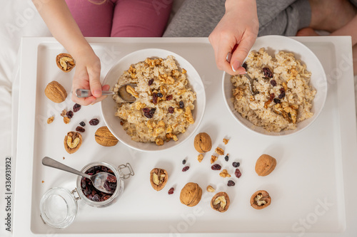 Happy mom and daughter have healthy breakfast on bed in a light bedroom on a sunny morning. Oatmeal porridge with nuts and raisins. Healthy food concept. Top view. Close-up
