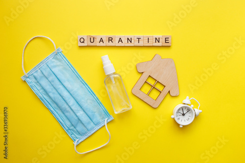 Top view of a yellow background with the word quarantine. Protective medical mask, alcohol spray and wooden house. Respiratory syndrome. World quarantine concept