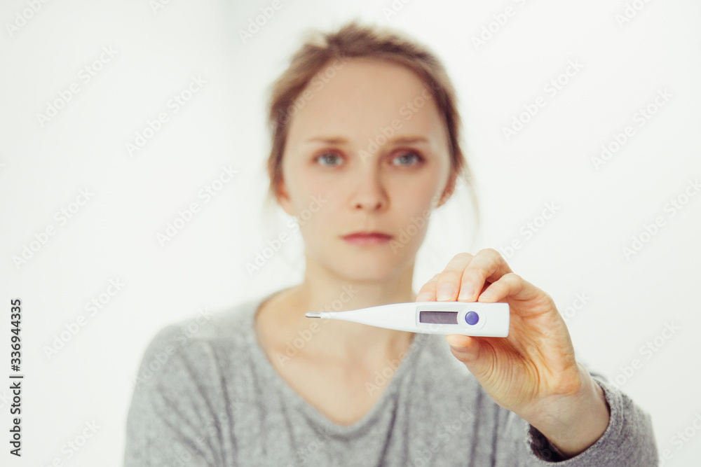 Sick young Caucasian woman in grey sweater sitting on bed, showing thermometre, looking at camera. Thermometre on focus. Illness, pain concept Sick young Caucasian woman in grey sweater sitting on bed, showing thermometre, looking at camera. Thermometre on focus. Illness, pain concept