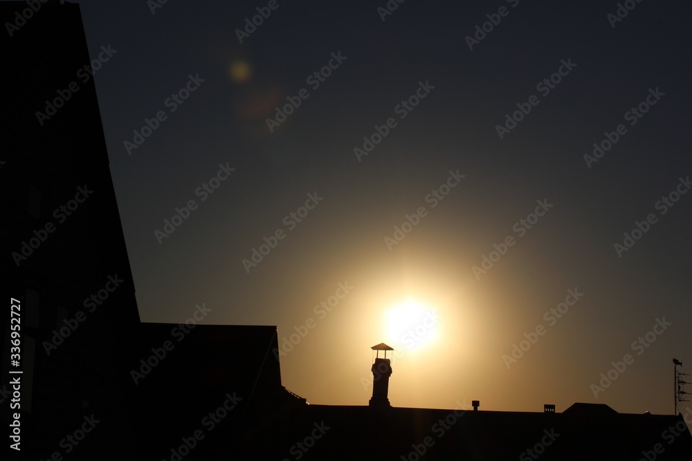 Sunset on the background. Shadow from the chimney against the background of the sun. row of house roofs, chimneys leading off to the horizon, silhouetted against a vivid sunset. Sunset on the background. Shadow from the chimney against the background of the sun. row of house roofs, chimneys leading off to the horizon, silhouetted against a vivid sunset.