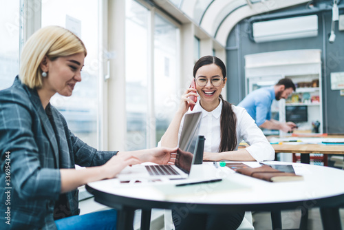 Cheerful woman chatting on phone sitting with female colleague using laptop