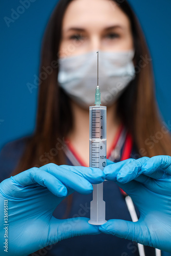 Portrait of caucasian female with ling dark hair in medical gown, blue medical gloves and special mask holds a syringe with medicine