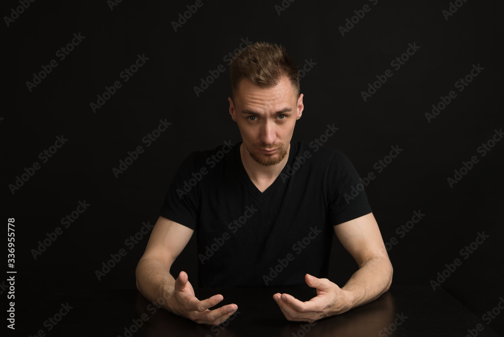 confused young man in a black t-shirt sits at a black table on a black background. put his hands on the table confused young man in a black t-shirt sits at a black table on a black background. put his hands on the table
