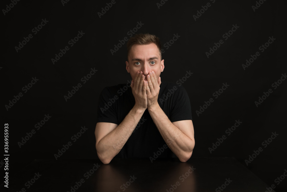 a man in a black shirt on a black background covers his mouth with his hands. Surprise