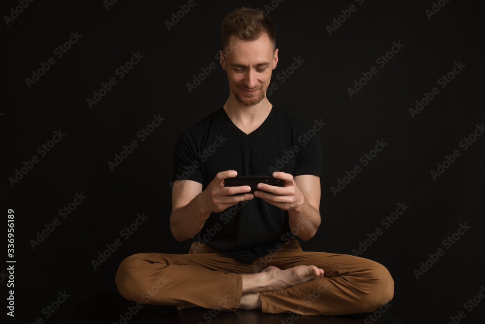 a man in a black T-shirt and brown pants on a black background sits in a lotus position and plays on the phone