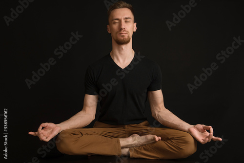a man in a black T-shirt and brown pants on a black background sits in a lotus position with his eyes closed