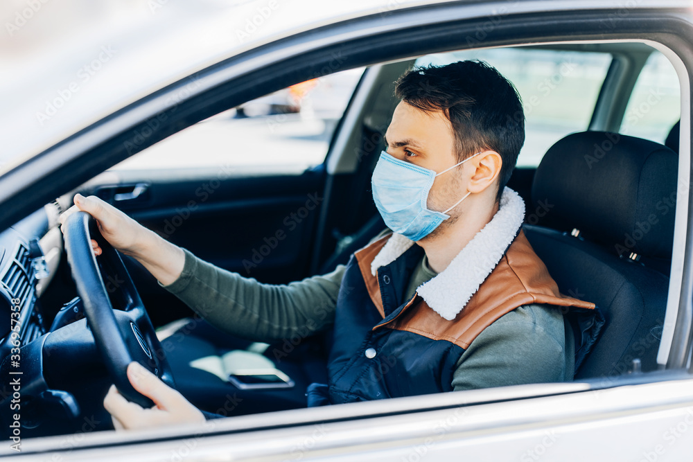 Beautiful young masked man sitting in a car, protective mask against coronavirus, driver on a city street during a coronavirus outbreak