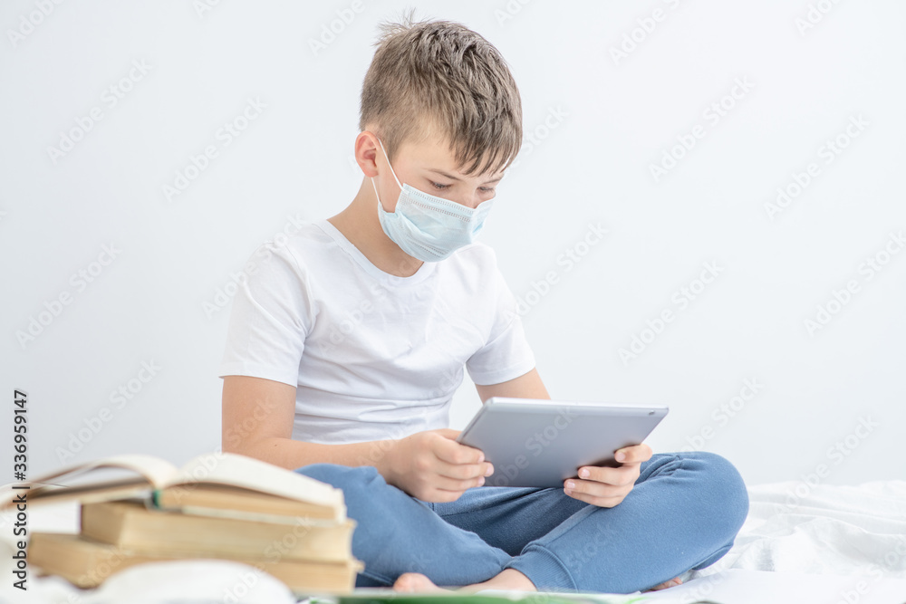 Smart boy wearing medical mask sits on the bed with tablet computer. Quarantine and coronavirus epidemic concept. Empty space for text Smart boy wearing medical mask sits on the bed with tablet computer. Quarantine and coronavirus epidemic concept. Empty space for text