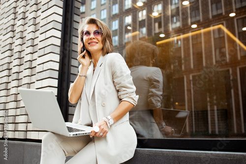 Stylish business lady working outdoor with her laptop. Freelancer working with pc in summer city. Fahionable female manager sit on the bench in the city park and typing.