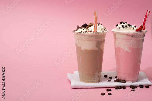 Disposable cups of chocolate and strawberry milkshakes with drinking straws and coffee grains on napkins on pink