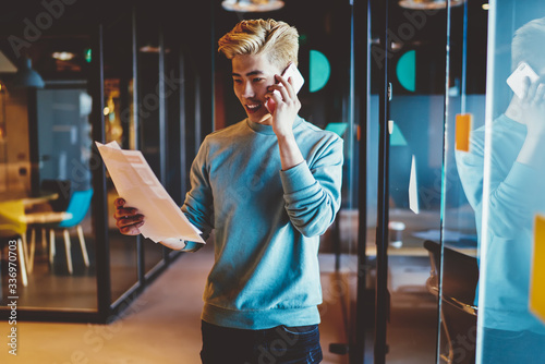 Cheerful blonde chinese male entrepreneur reading document and having mobile phone conversation in office, skilled asian businessman making banking via smartphone call holding papers with transactions