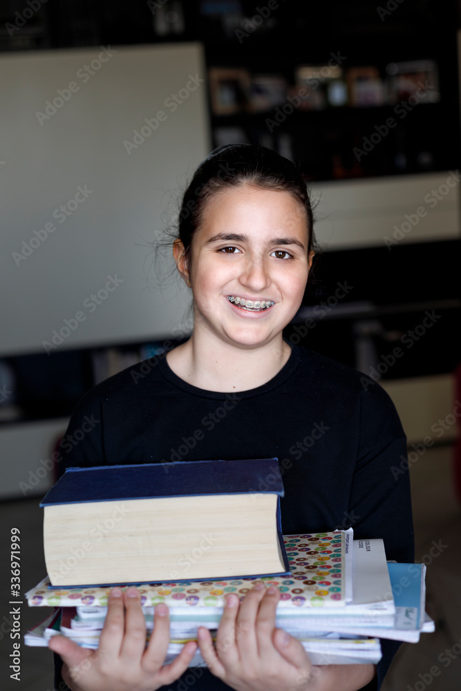 Fototapeta premium Ritratto di giovane agazza coni capelli scuri raccolti e un maglia nera che tiene in mano tanti libri per studiare, isolata su sfondo nero