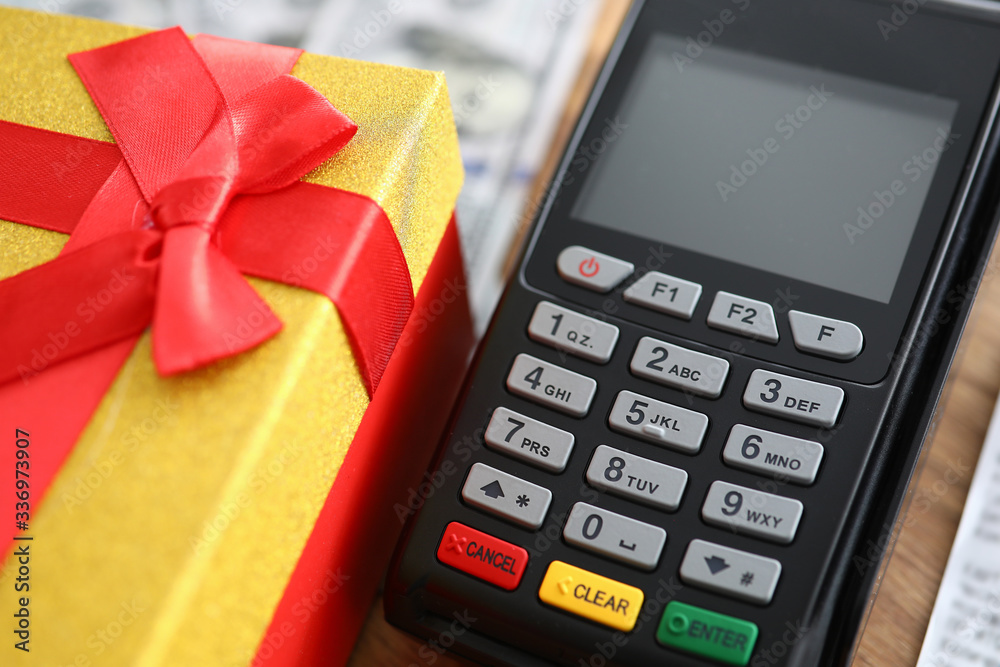 Close-up of terminal machine and yellow present box with red bow on top. Spending money on online shopping. Pay via credit card. Modern technology concept Close-up of terminal machine and yellow present box with red bow on top. Spending money on online shopping. Pay via credit card. Modern technology concept