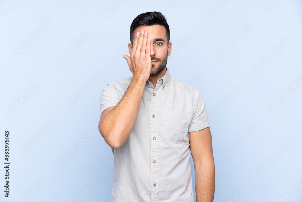 Young handsome man with beard over isolated blue background covering a eye by hand Young handsome man with beard over isolated blue background covering a eye by hand