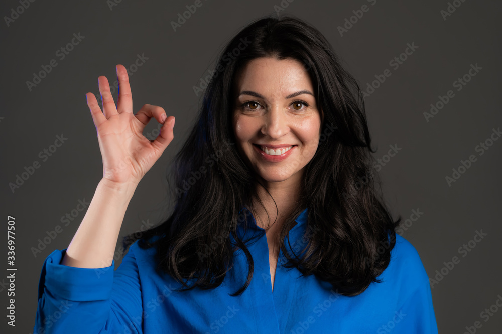 Happy adult woman on grey background. Lady with long hair in blue shirt shows Ok gesture. Winner. Success. Positive lady smiles to camera. Body language. Happy adult woman on grey background. Lady with long hair in blue shirt shows Ok gesture. Winner. Success. Positive lady smiles to camera. Body language.