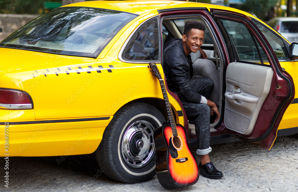 Horizontal view. Smiling young man in interior salon of car with openend door of yellow car, looking a side, with left leg outside, near guitar. Horizontal view. Smiling young man in interior salon of car with openend door of yellow car, looking a side, with left leg outside, near guitar.