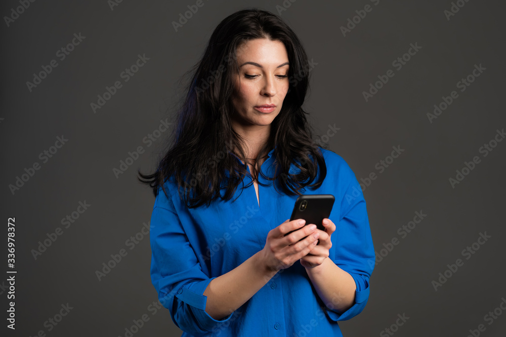 Mature woman sms texting, using app on smartphone. Pretty adult lady surfing internet with mobile phone. Grey studio portrait. Mature woman sms texting, using app on smartphone. Pretty adult lady surfing internet with mobile phone. Grey studio portrait.