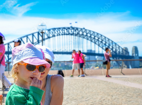 Little girl in cap and sunglasses with view of Sydney Harbor Bridge from Sydney Opera House square, the girl is 3 years old and eats a small snek