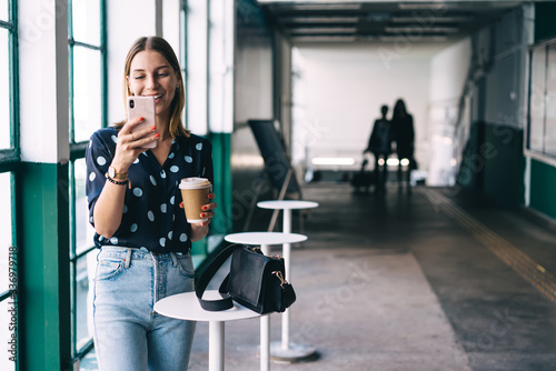 Happy caucasian blonde hipster girl reading message from social networks on mobile phone during coffee break, positive 20s woman in trendy wear watching videos and chatting via smartphone and 4G