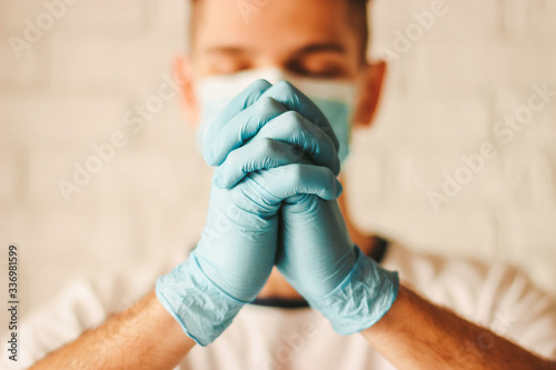 Religious catholic doctor in medical face mask and protective gloves pray to God in hospital. Young man christian prayer in medical gloves and protective face mask in church. COVID-19 coronavirus help