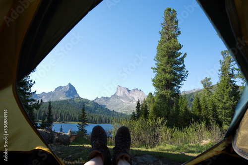Inside view of the tent on a beautiful lake surrounded by mountains, tall trees and pine trees grow in front of the tent. Traveler lie in a tent overlooking a mountain landscape in Russia